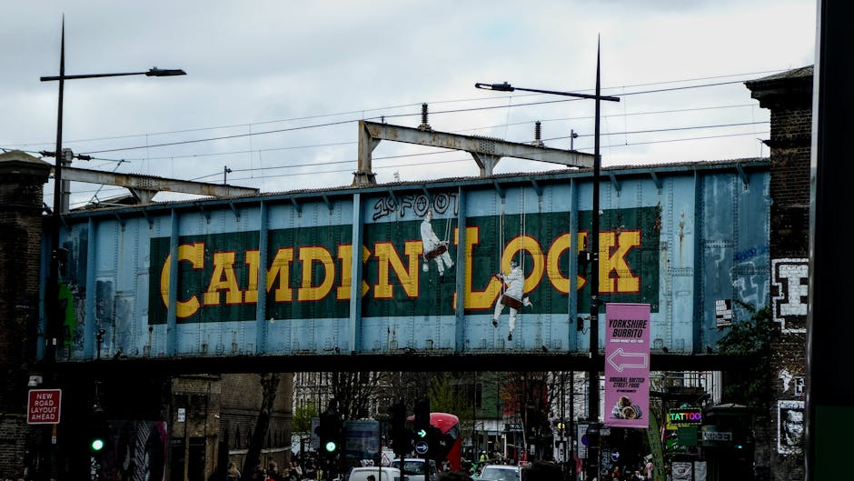 A street scene outside Camden Lock Market in London, featuring a large, green and yellow sign with the words 'Camden Lock' mounted on an elevated structure above the road. In the foreground, there are